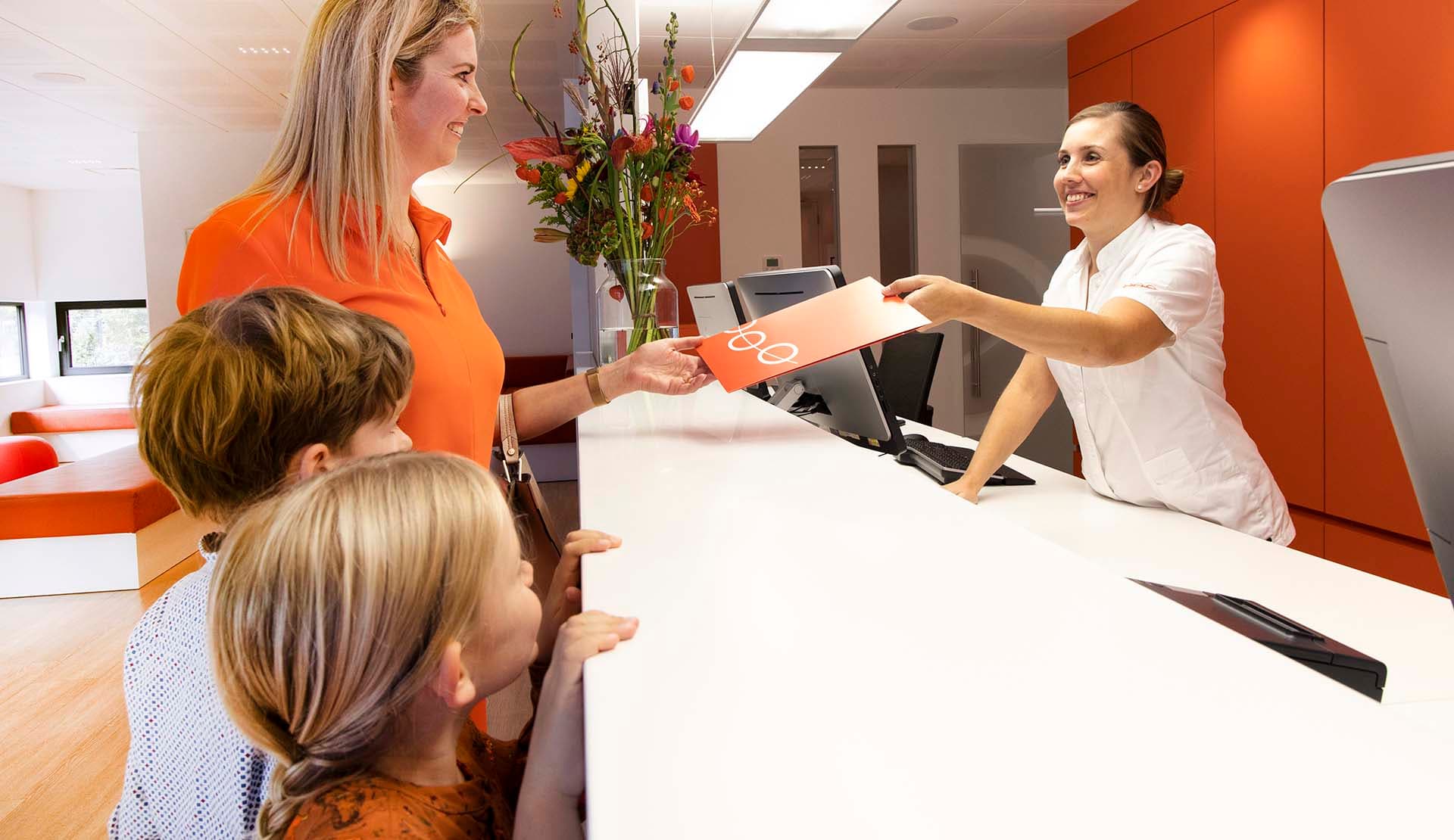Een patiënt staat bij de balie van een orthodontist met twee kinderen. Achter de balie staat een vrouw die hen helpt.
